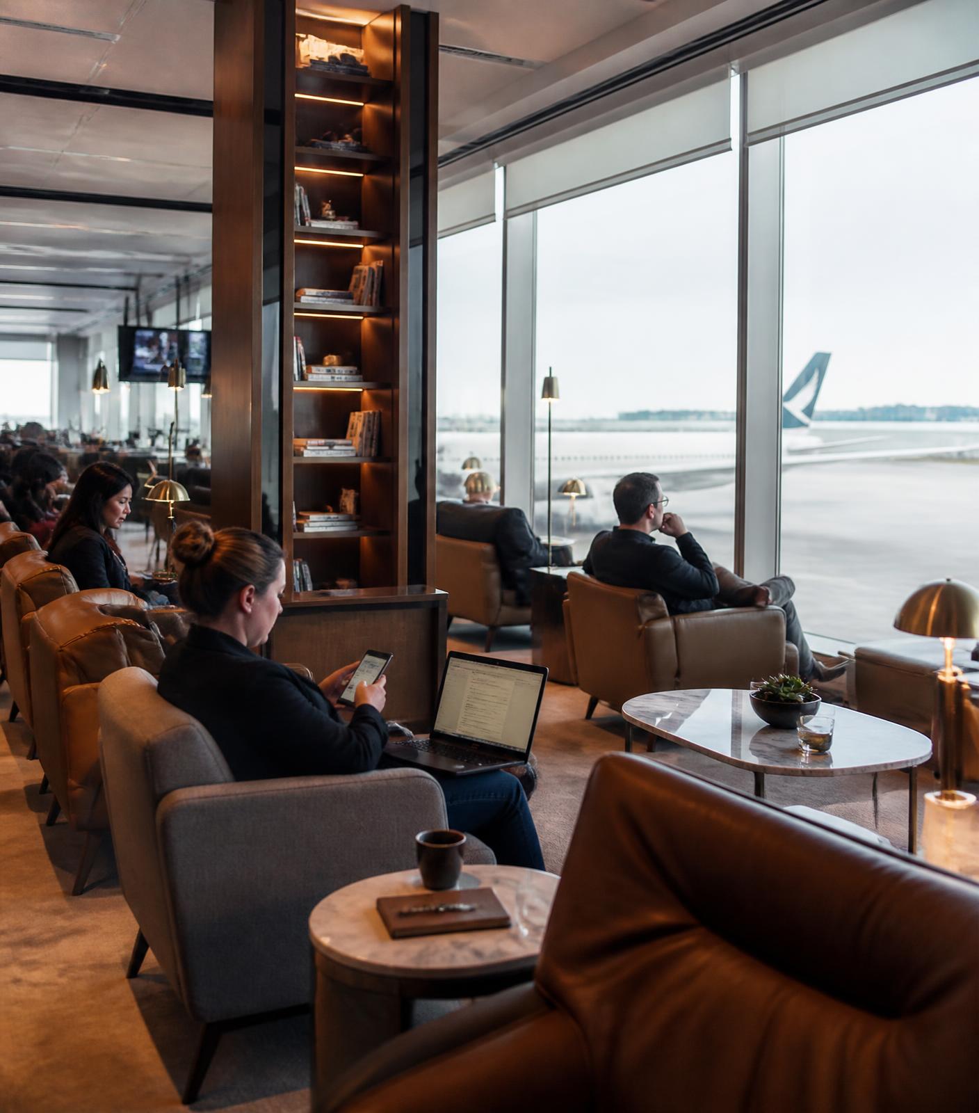 Modern airport lounge with comfortable seating and travellers relaxing beside floor-to-ceiling windows overlooking the apron